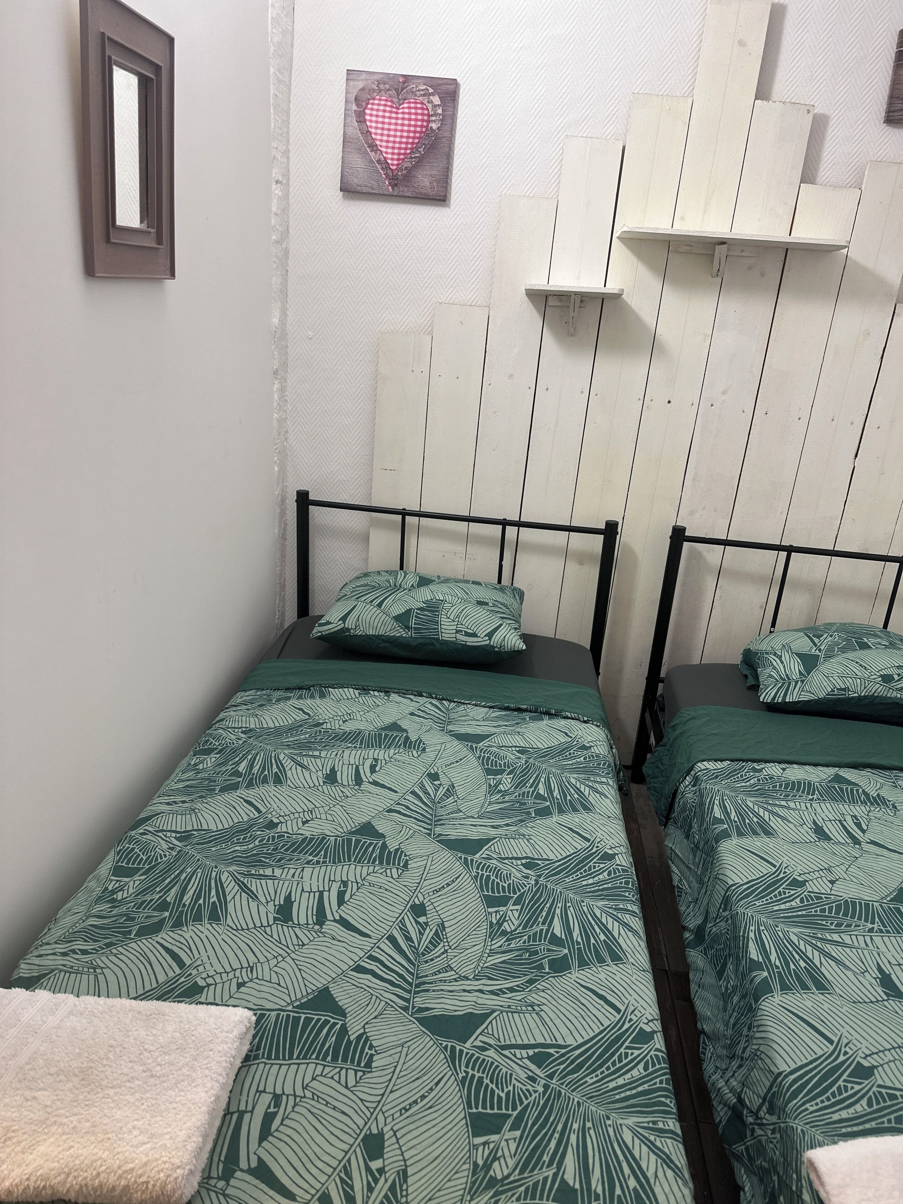 A room with two single beds, each with a green leaf-patterned bedspread and matching pillows. There is a small heart-shaped decoration on the wall above the beds.