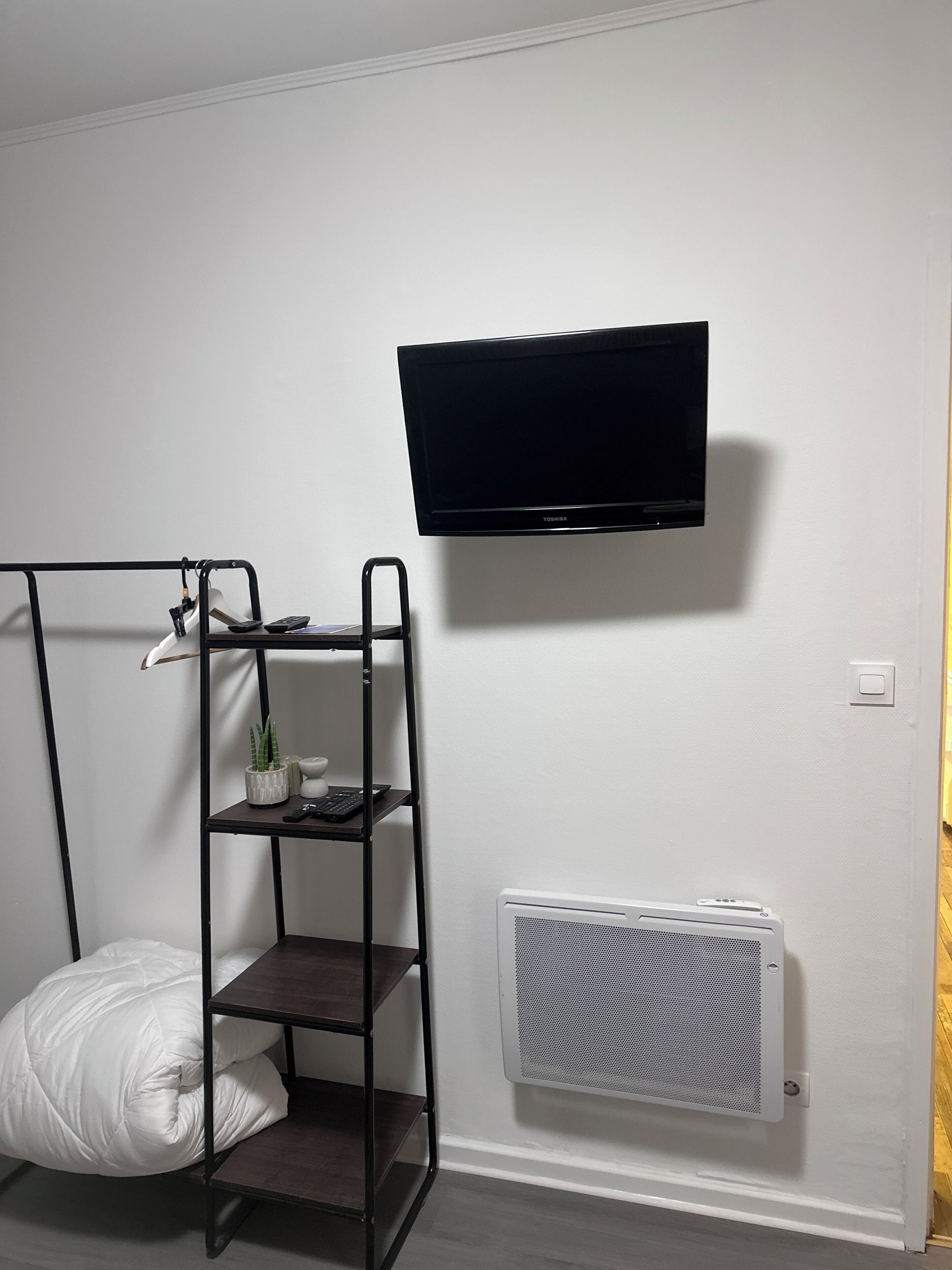 A room with a bed, a TV mounted on the wall, a radiator, and a shelf with a plant and a keyboard.