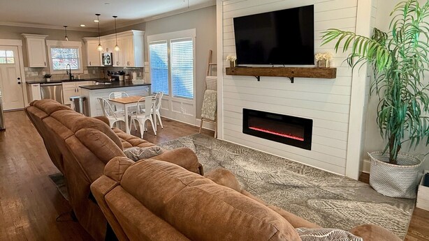 A modern living room with a large sectional sofa, a fireplace, a flat-screen TV mounted on the wall, and a kitchen in the background.