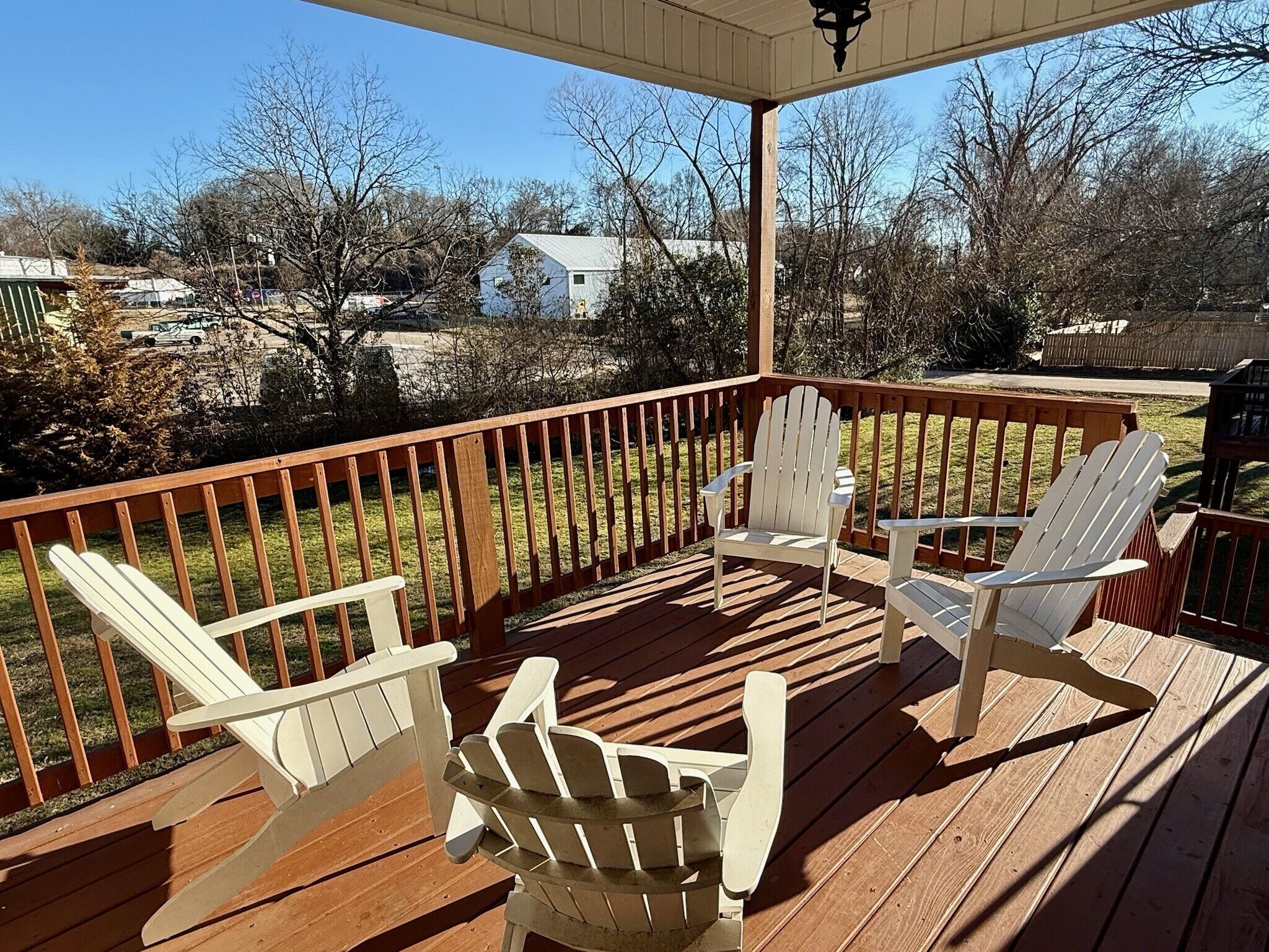Une terrasse en bois avec trois chaises Adirondack blanches, un espace abrité avec une rambarde, et une vue sur des maisons et des arbres en arrière-plan.