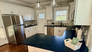 A modern kitchen with white cabinets, a stainless steel refrigerator, and a dark countertop. There are two bar stools and a basket with a book on the counter.