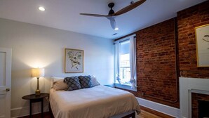 A bedroom with a bed, a nightstand, a lamp, a ceiling fan, a window with curtains, a brick wall, and a fireplace. - The Nest Above - Finch Suite (Seneca)