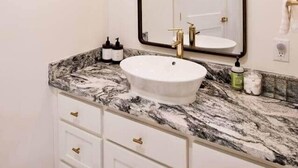 A bathroom with a white sink, marble countertop, gold faucets, and a large mirror. - The Nest Above - Finch Suite (Seneca)