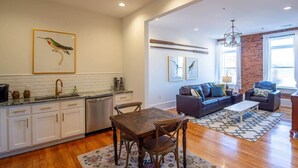 A spacious living area with a wooden dining table, a sofa set, and a kitchen with white cabinets and a dishwasher. - The Nest Above - Finch Suite (Seneca)
