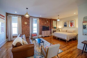 A modern living room with a brick wall, a sofa, a TV, a fireplace, and a bed. - The Nest Above -Bluebird (studio) (Seneca)
