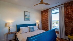 Un dormitori amb un panell blau al capçal, un finestral gran amb cortines, un ventilador de sostre i un quadre a la paret.
