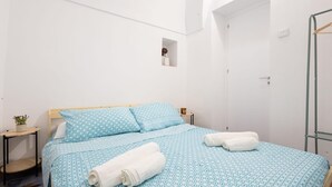 Ein Zimmer mit einem Einzelbett, einer blauen Bettdecke mit Muster, zwei Kissen, einem Holzkopfteil, einem Nachttisch mit Pflanze und einem kleinen Spiegel an der Wand.