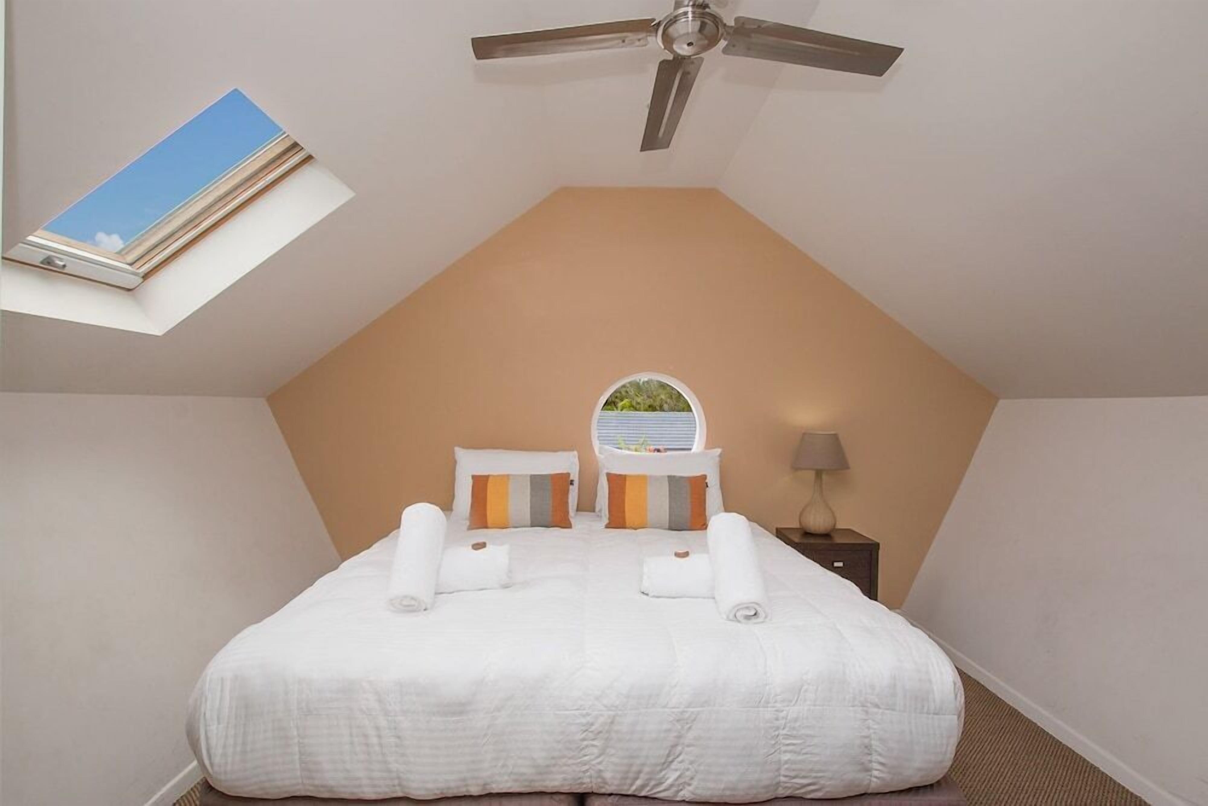 A neatly made bed with white linens and rolled towels, a ceiling fan, a skylight, and a small bedside table with a lamp.