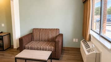En stue med sofa, et lille bord, et vindue med gardiner og en vægmonteret aircondition.