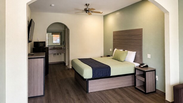 Quarto com cama, mesa de cabeceira, escrivaninha com cadeira, ventilador de teto e uma porta arqueada que dá para um banheiro.