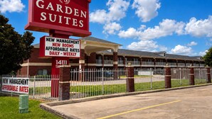 Placa de hotel com os dizeres 'Garden Suites' e 'Nova Administração', um edifício de tijolos com cerca branca e um céu azul claro.
