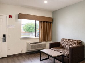 A hotel room with a single sofa, a small table, a window with curtains, a wall-mounted air conditioner, and an emergency exit sign. - GARDEN SUITES (Houston)