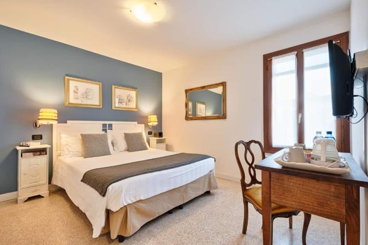 A bedroom with a bed, bedside tables, a desk with a chair, a mirror, and artwork on the wall.