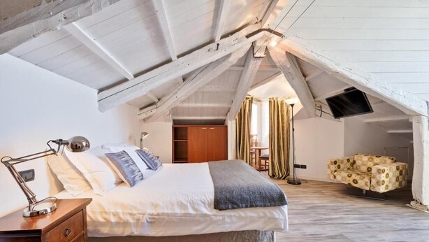 Une chambre avec un plafond en pente, un lit avec des oreillers, une table de chevet, un téléviseur fixé au mur et un canapé à motifs.