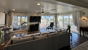 A spacious living room with a large sofa, a flat-screen TV, and a balcony with a view. - IR56: Beach Belle: Serene Marsh Views (Ocracoke)