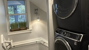 A laundry room with a large black washer and dryer, a window with a view of trees, and a small potted plant on the windowsill. - IR56: Beach Belle: Serene Marsh Views (Ocracoke)