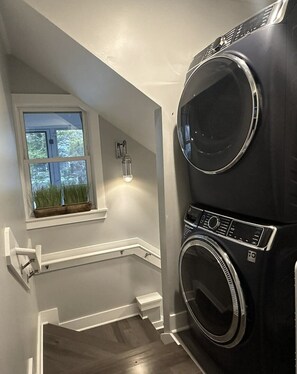 A laundry room with a large black washer and dryer, a window with a view of trees, and a small potted plant on the windowsill. - IR56: Beach Belle: Serene Marsh Views (Ocracoke)