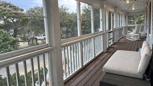 A covered porch with a wooden deck, white railings, and a white cushioned bench. - IR56: Beach Belle: Serene Marsh Views (Ocracoke)