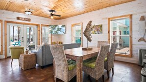 A cozy cabin interior with a wooden ceiling, a dining area with wicker chairs, a large TV, and a view of the beach through the windows.