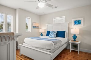 Un dormitorio con una cama blanca, almohadas azules, dos mesitas de noche con lámparas y un ventilador de techo.
