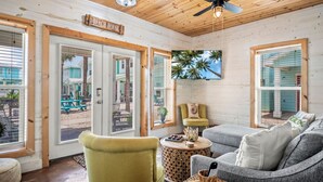 A beach house interior with a ceiling fan, a sign reading "BEACH HOUSE", and a view of the beach through large windows.