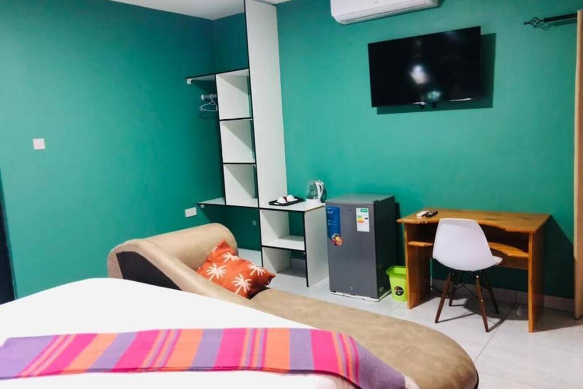 Une pièce aux murs de couleur turquoise, avec un lit, un bureau, une chaise, un téléviseur et un réfrigérateur.