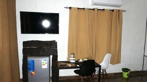A room with a TV mounted on the wall, a small refrigerator, a table with a chair, and a window with curtains. - Makgadikgadi Riverview Inn (Nata)