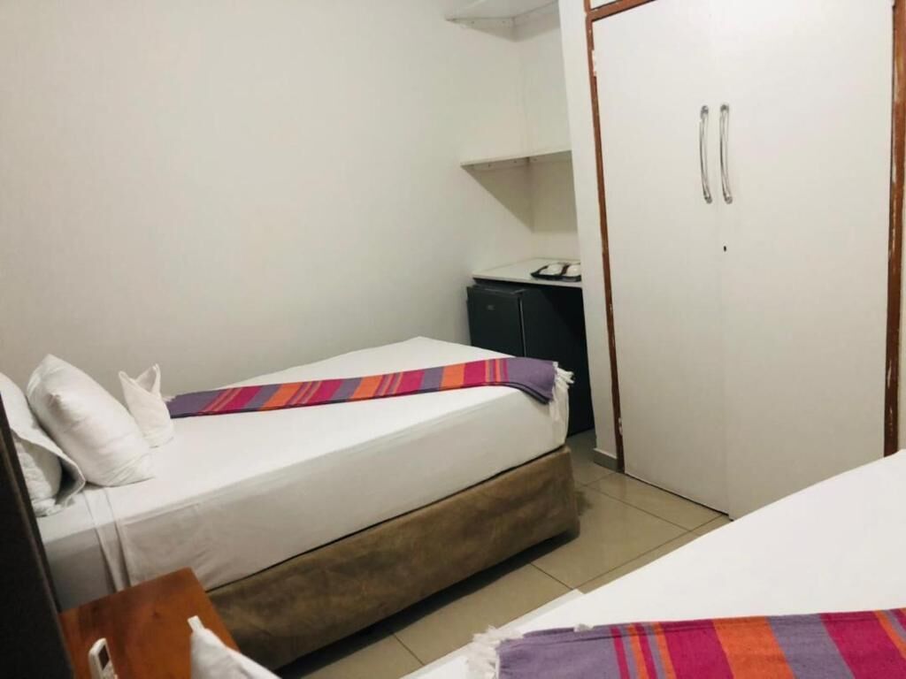Una habitación pequeña con cama individual, estructura de madera, colchón blanco y manta colorida. Hay un escritorio pequeño con una computadora y un armario con espejo. Las paredes son blancas y hay un guardarropa de madera con dos puertas.