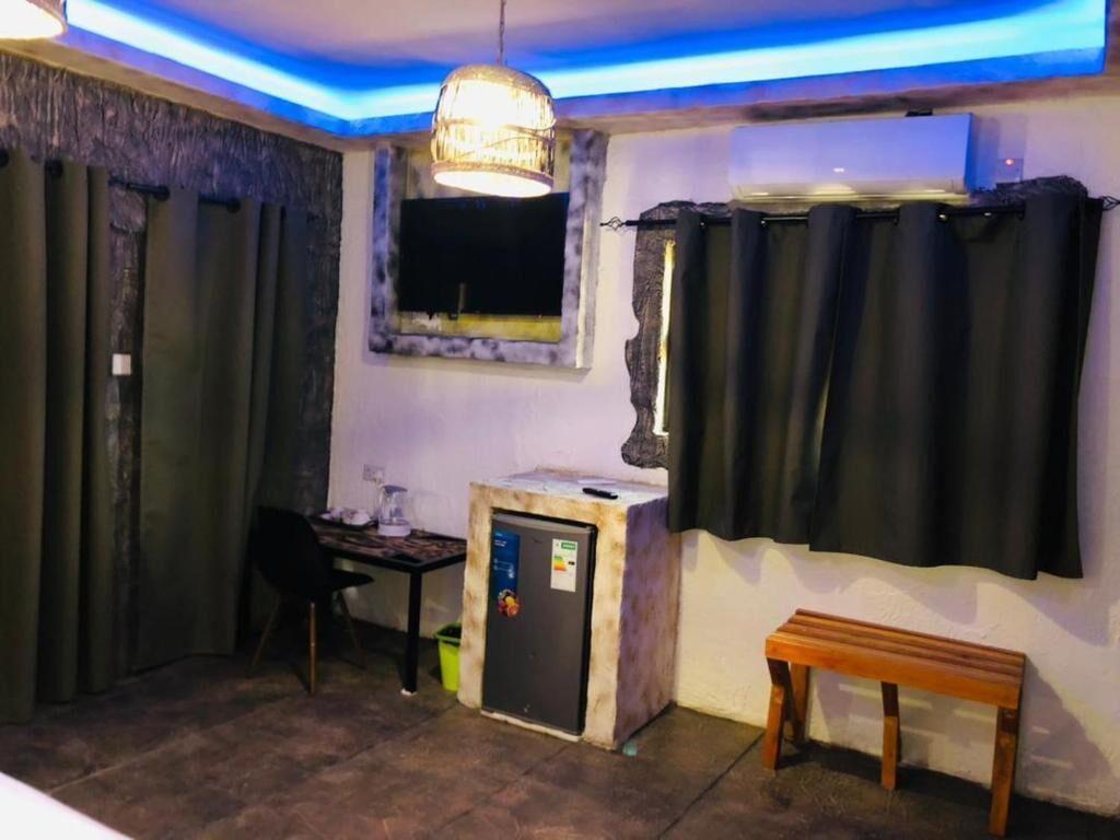 A small room with a desk, a chair, a small refrigerator, a bench, and a window with curtains.