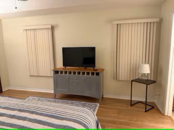 Un dormitorio con cama, televisor en un mueble, mesita de noche con lámpara y dos ventanas con cortinas.