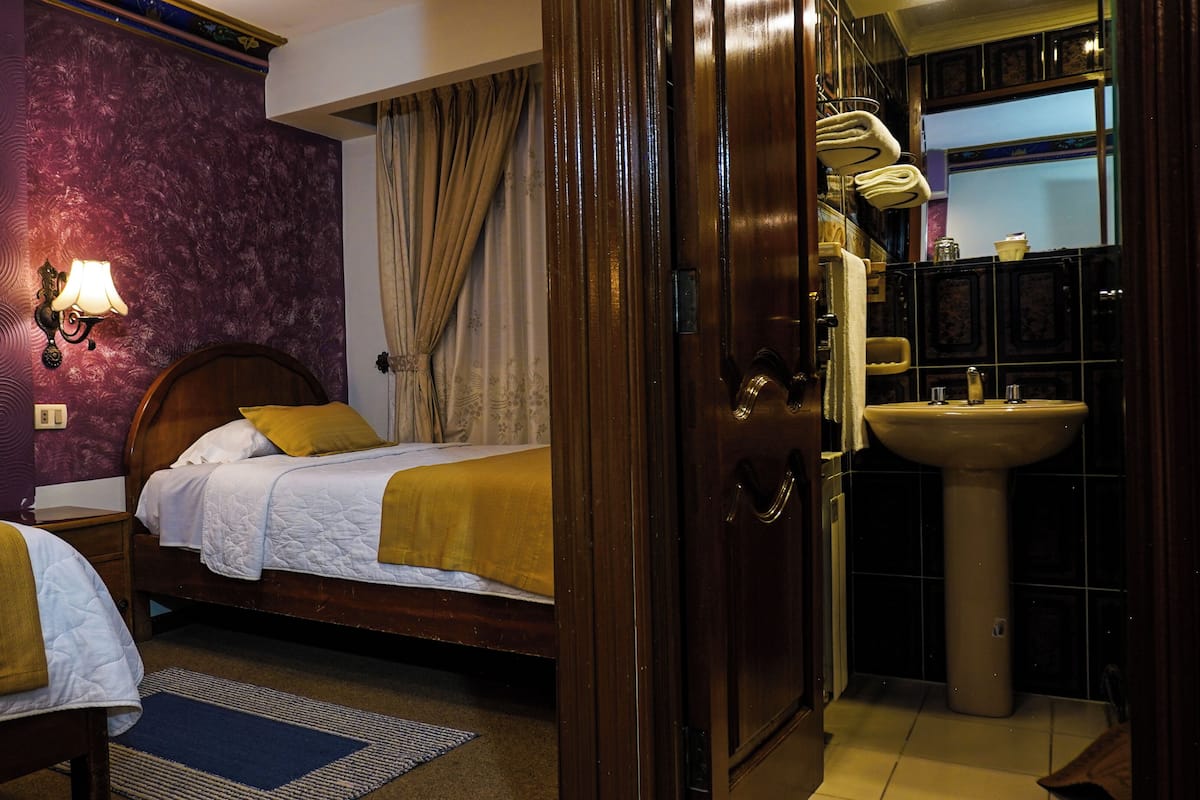 Habitación de hotel con dos camas, cabecero de madera, mesita de noche con lámpara, baño con lavabo y espejo, y ventana con cortinas.
