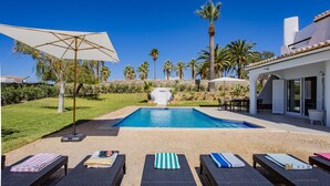 A pool area with lounge chairs, a covered seating area, and palm trees. - Greice Homes - Villa Carob Tree (Almancil)