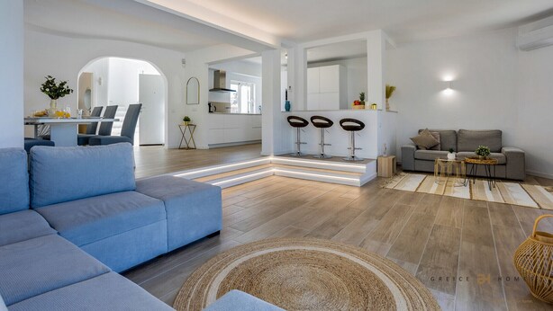 A modern living room with a sectional sofa, a dining area with a table and chairs, a kitchen with bar stools, and a grey sofa in the living area. - Greice Homes - Villa Carob Tree (Almancil)