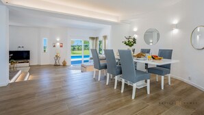 A modern dining area with a white table, grey chairs, and a view of a swimming pool. - Greice Homes - Villa Carob Tree (Almancil)