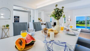 A modern dining area with a table set for breakfast, featuring fruits, bread, and beverages. - Greice Homes - Villa Carob Tree (Almancil)