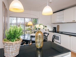 A modern kitchen with white cabinets, a black countertop, and two pendant lights. There is a dining table with chairs, a vase with flowers, and a potted plant. - 4 Star Holiday Home in Tylstrup (Tylstrup)