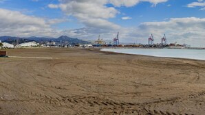 Una playa amplia de arena con un mar tranquilo, montañas a lo lejos y grúas industriales.