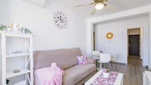 Una sala de estar moderna con un sofá beige, una mesa de centro blanca con camino de mesa floral, una estantería blanca y un reloj de pared.