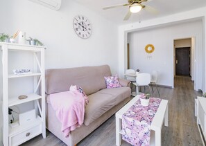 Ein modernes Wohnzimmer mit einem beigefarbenen Sofa, einem weißen Couchtisch mit floralem Tischläufer, einem weißen Bücherregal und einer Wanduhr.