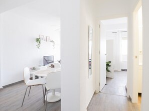 Ein moderner, minimalistischer Raum mit weißem Schreibtisch, Stuhl und Computer. An der Wand befinden sich gerahmte Bilder und ein kleiner Topf mit Pflanze.