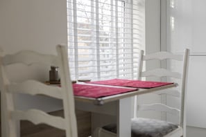 A white chair with a cushion, a table with a pink placemat, and a window with blinds. - Immaculate Apartment | Historic Lincoln | Wi-Fi (Lincoln)