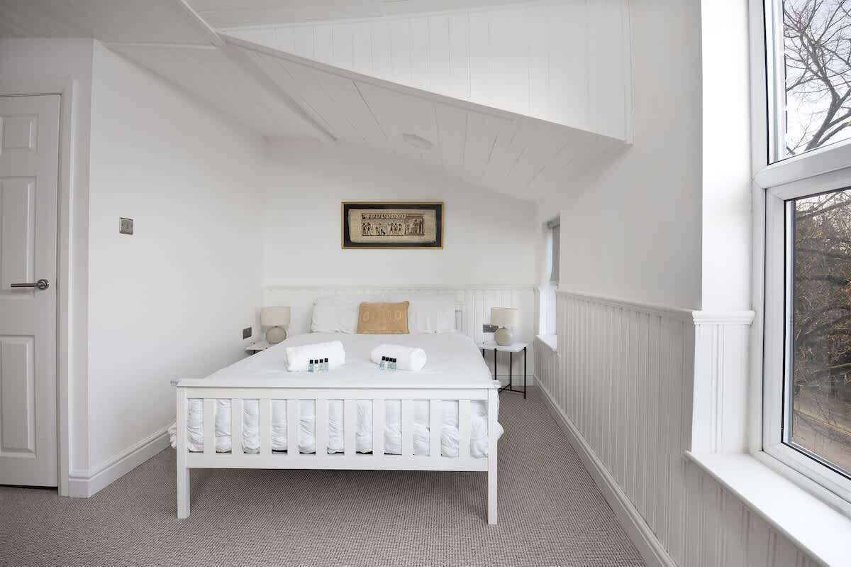 Serene bedroom with crisp white walls, a comfy double bed, and a cozy lounge chair for reading or relaxing. Enjoy your favorite shows on the TV, and let natural light fill the space through the window, creating a peaceful retreat after a busy day.