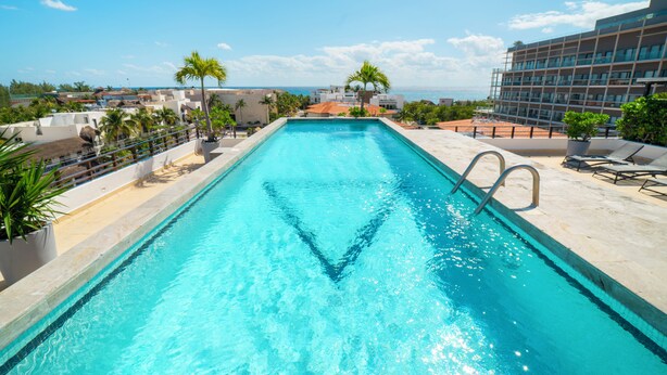Ein rechteckiger Swimmingpool mit klarem, blauem Wasser, umgeben von einer weißen Poolterrasse, Palmen und im Hintergrund Blick auf das Meer und Gebäude.