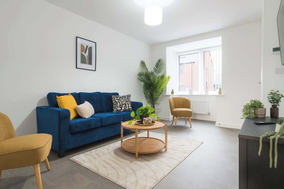 A modern living room with a blue sofa, a round wooden coffee table, a yellow armchair, and a large window with blinds.