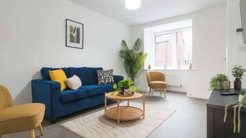 A modern living room with a blue sofa, a round wooden coffee table, a yellow armchair, and a large window with blinds.