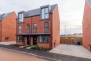 Une maison moderne à deux étages, construite en briques, avec un porche à l’avant, une cour arrière clôturée et un ciel dégagé.