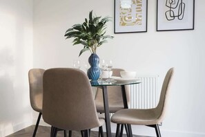 Un coin repas avec une table ronde en verre, deux chaises beiges, un vase bleu contenant des plantes vertes et des œuvres d’art encadrées accrochées au mur.