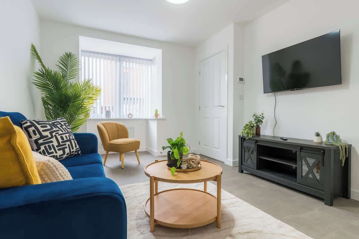 A living room with a blue sofa, a round wooden coffee table, a television on the wall, and a large window with blinds.