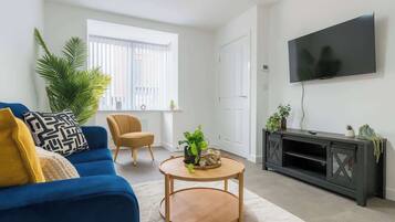 A living room with a blue sofa, a round wooden coffee table, a television on the wall, and a large window with blinds.