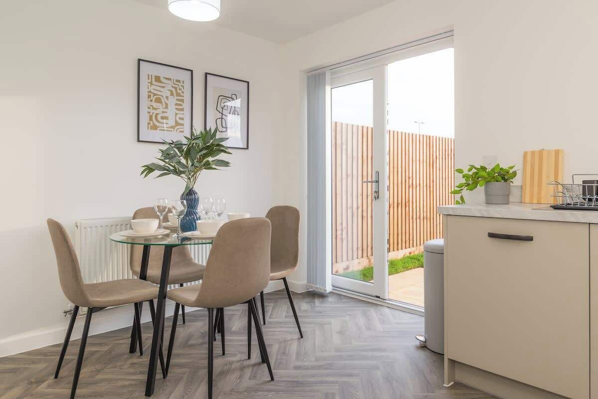 A dining area with a glass table, four chairs, a vase with a plant, and two framed artworks on the wall.